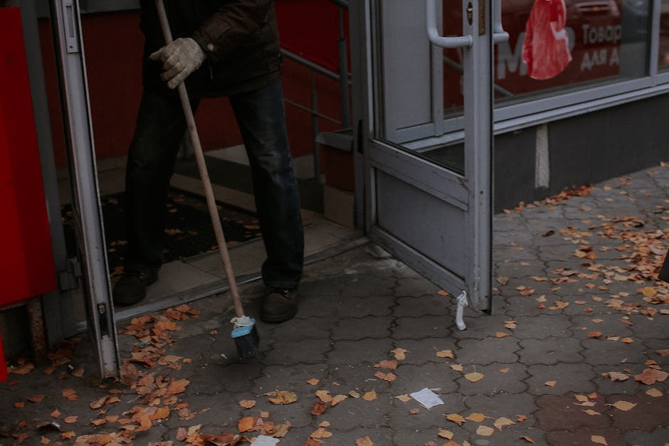 Person sweeping dry leaves outside a shop entrance during fall.