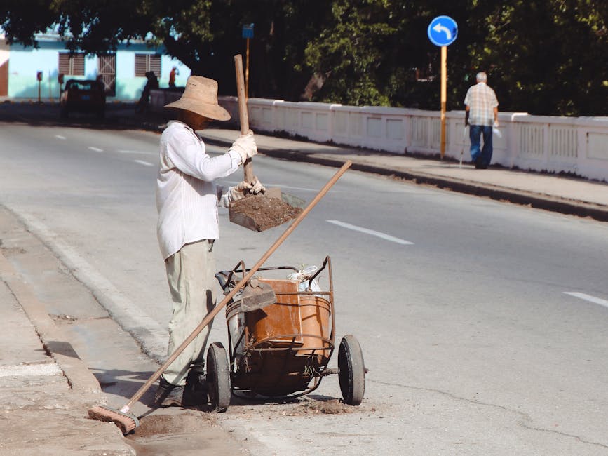 A person sweeping the street with a broom and a cart in an urban setting.